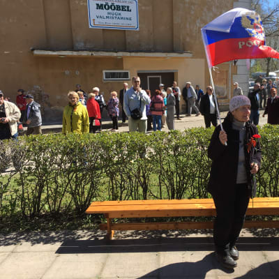 Mustiin vaatteisiin pukeutunut vanhempi nainen pitää punaisia neilikoita ja heiluttaa Venäjän lippua aurinkoisena kevätpäivänä virolaisessa Kohtla-Järven kaupungissa. 