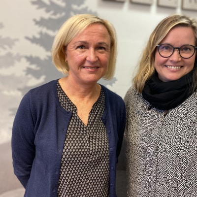 Lähikuvassa kaksi vaaleahkohiuksista naista, vasemmalla polkkatukkainen nainen sinisessä villatakissa ja oikealla hiukan nuorempi silmälasipäinen ja pitkätukkainen nainen. Molemmat hymyilevät. 