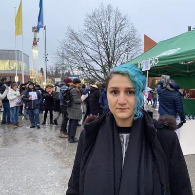 Aalto-yliopistossa opiskeleva Fereshteh Sohrabi katsoo kameraan Espoon Otaniemessä ja taustalla näkyy Iranin kansan puolesta järjestetyn mielenosoituksen osallistujia.