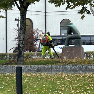 Työhaalareihin sonnustautunut henkilö käyttää lehtipuhallinta patsaan vieressä.