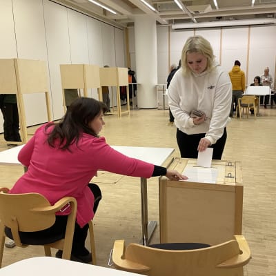 Nainen istuu selin pöydän ääressä pinkki jakku päällä ja auttaa kasvot kameraan päin olevaa vaaleaan huppariin pukeutunutta naista pudottamaan äänestyslipukkeen uurnaan.
