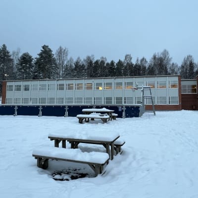 Lumen peittämä koulupiha, jossa on muutamia puisia penkkejä ja pöytiä. Taustalla kaksikerroksinen tiilitalo.