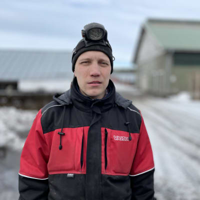 En man i arbetsoverall står ute på en pälsfarm med mössa och pannlampa i snömodden. 
