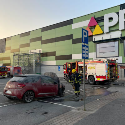 Palanut auto Prisman pihassa. Taustalla paloauto ja palomiehiä.