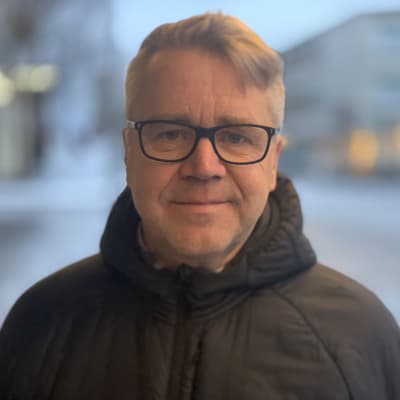 En man med grått hår och mörka bågar på glasögonen står i en urban miljö med vinterjacka. 