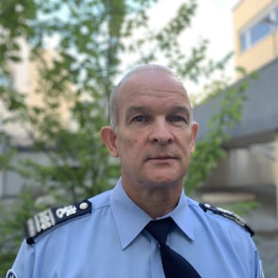 Mansperson i polisuniform står utomhus, i bakgrunden sommargrönska.