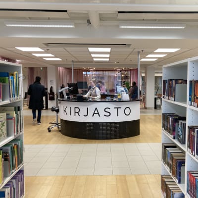 KIrjahyllyjä ja palvelutiski, jossa lukee KIRJASTO. Taustalla asiakkaita ja kirjastotyäntekijä.