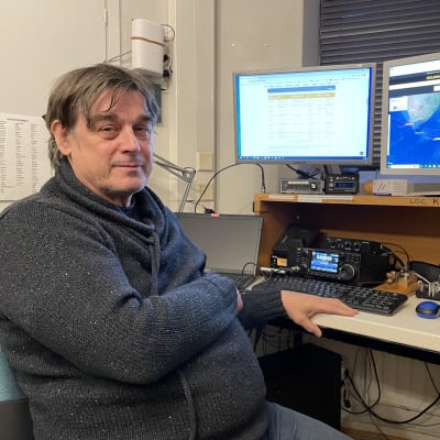 Jari Jussila istuu pöydän ääressä. Taustalla radiolaitteita ja kaksi tietokoneen näyttöä.