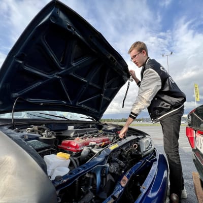 Nuori silmälasipäinen mies on avannut sinisen Nissan-merkkisen auton konepellin ja näyttää moottoria.