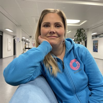 Nainen istuu sohvalla, nojaa kasvojaan käteensä ja katsoo hymyillen kameraan.