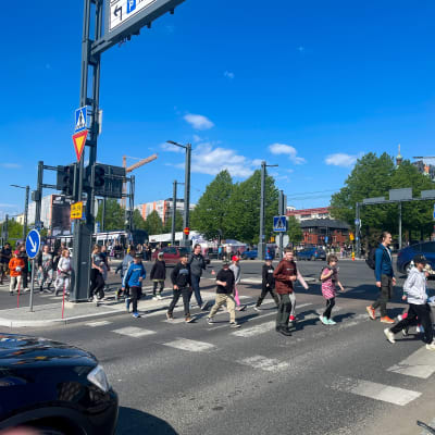 Lapsia ja nuoria ylittämässä suojatietä Tampereen keskustassa. Aurinko paistaa kirkkaalta taivaalta.