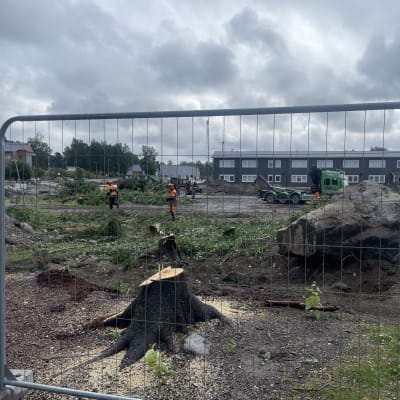 En byggarbetsplats med två personer klädda i orangea arbetskläder. Bakom dem finns en grön lastbil och en röd traktor.