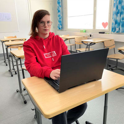 Sysmän lukion opiskelija Lumi Renvall punaisessa hupparissa pulpetin ääressä tietokone edessään.