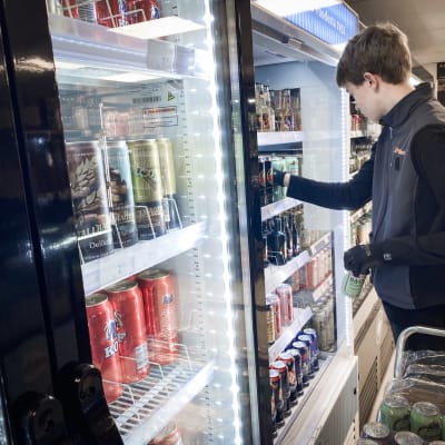 En butiksanställd fyller på ett kylskåp med öl- och läskburkar i en matbutik.