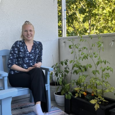 Nuori nainen istumassa puisella parveke-tuolilla parvekkeellaan. Parvekkeella erilaisia kukkaruukkuja ja -penkkejä, joissa hyötykasveja.