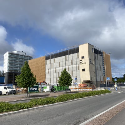 Iso uusi rakennus, jossa on ruskeaa puuverhoilua ja vaaleaa seinää. Rakennuken kyljessä luke P Kartano. Vasemmalla näkyy kerrostalo. Rakennusten edessä on autoja ja ohite kulkee tie, jonka keskellä on pensaita.