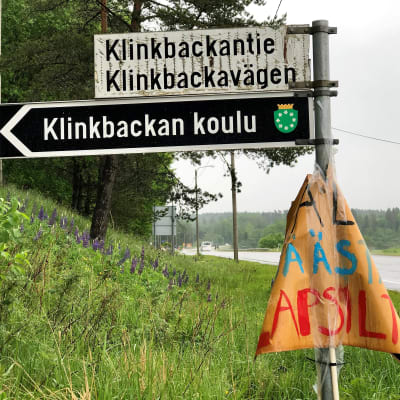 En vägskylt med finsk och svensk text. Nedanför vägnamnen står det på en svartvit skylt Klinkbackan koulu. Till höger löper en asfaltväg. Regnig väder. Grön försommarmiljö.