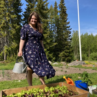 Maria Haglund står bakom sin pallkrage med sallad som hon odlat. Hon har en klänning på sig och håller i en vattenkanna.