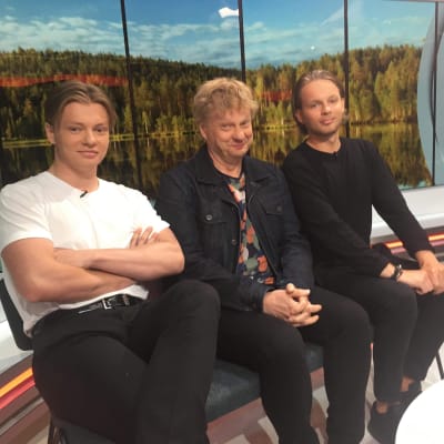 Iiro Rantala poikiensa kanssa istumassa tv-studiossa, taustalla on kuva suomalaisesta järvimaisemasta. 