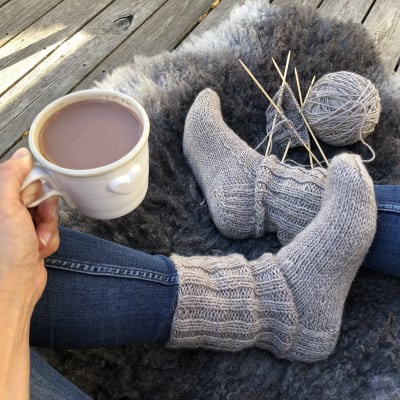 Fötter iklädda stickade sockor, stickning och garnnystan på fårfäll. En hand som håller en kopp varm choklad.