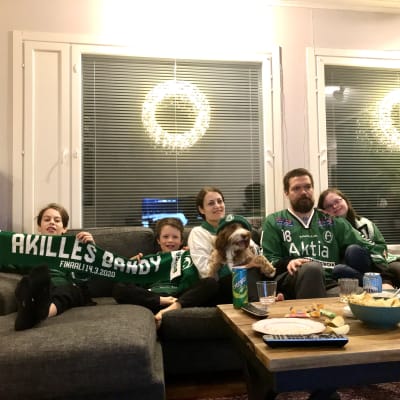 En familj med mamma, pappa och tre barn sitter i en soffa med en hund. De har gröna mössor och halsdukar på sig.