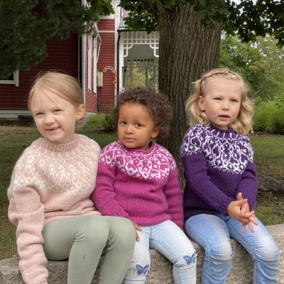 Kolme tyttöä istuu penkillä. Käsinneulotut strömsö lastenvillapaitoja pukeutuneena.