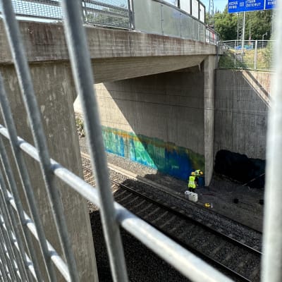 Två västklädda män målar över graffiti intill en järnväg.
