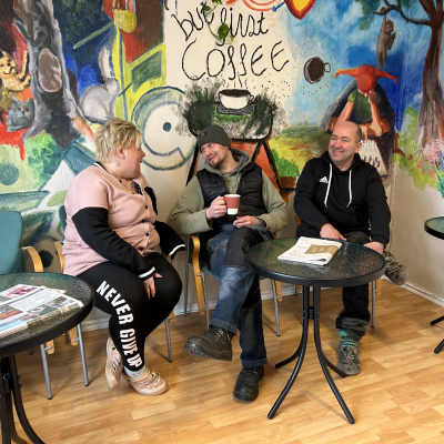 Kafferummet i Klubbhus Fontana i Karis. På väggen en stor väggmålning i granna färger. Runt bordet sitter Linda Färm från Pojo, Samuli Lindholm från Antskog och Christian Karén från Karis.