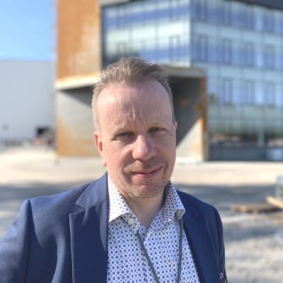 Mansperson i blå skjorta och kavaj står framför nybyggd kontorsbyggnad.