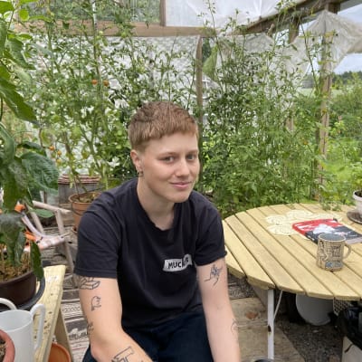 Edith Hammar sitter i sitt växthus och tittar in i kameran. I bakfgrunden skymtar växter och gröna kvistar, och boken "Homo Line" på bordet. 