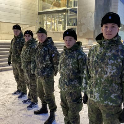 Viisi nuorta asevelvollista miestä seisoo rivissä varuskunnan lumisella pihalla esikuntarakennuksen edessä aamuhämärässä.