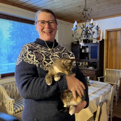 Marika Rinta-Tassi seisoo olohuoneessa, hymyilee ja pitää kissaa sylissään.