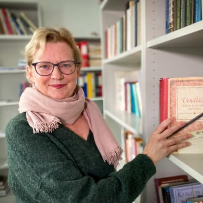 Sanna Turoma, Venäjän kielen ja kulttuurin professori, Tampereen yliopistolla, päällään vihreä villapaita ja kaulassa vaaleanpunainen kaulahuivi. Seisoo kirjahyllyn vierellä, kädessään Venäjää käsittelevä kirja. 