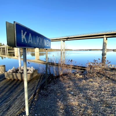 Kalkkisen kylän kyltti Kymijoen rannalla kirkkaana kevätaamuna kuvattuna.