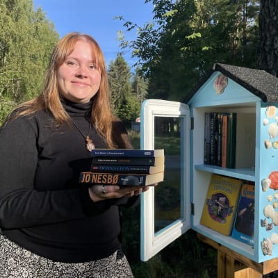 Muhoslainen Mirette Piekkari esittelee tienlaidassa sijaitsevaa minikirjastoa, joka muistuttaa lasiovista linnunpönttöä.