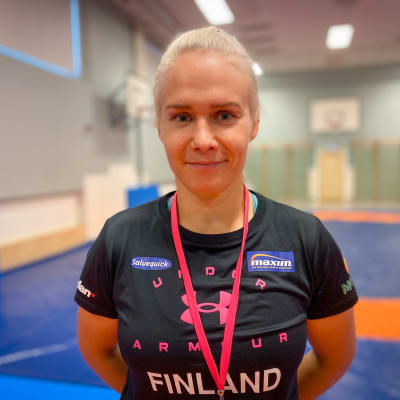 Vaaleahiuksinen, mustaan t-paitaan pukeutunut Petra Olli seisoo urheilusalissa kädet selkänsä takana ja hymyilee kameralle.