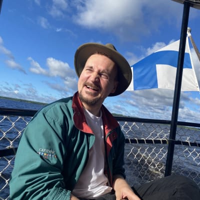 Mies hymyilee laivan kannella, Suomen lippu liehuu taustalla.