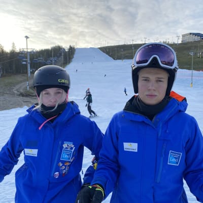 Alppikoululaiset Peppi Ravolainen ja Altti Pyrrö sinisissä takeissa ja laskettelukypärissä katsovat kameraan, taustalla Levin säilölumirinne.