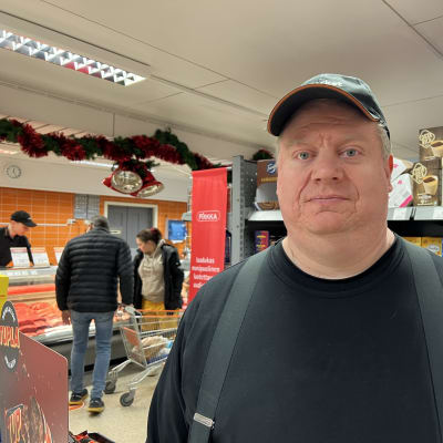 Kauppias Juha-Matti Lias seisoo kaupan sisätiloissa, taustalla kaksi asiakasta ja lihatiskin myyjä.