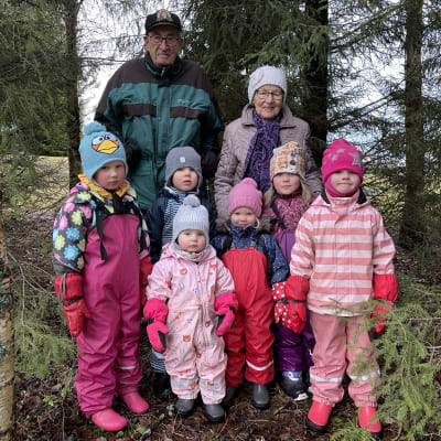 Metsämummo, metsäukki ja kuusi lasta poseeraavat kameralle metsässä. 