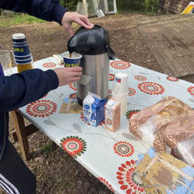 En persons händer syns som tar kaffe i en kaffetermos utomhus.