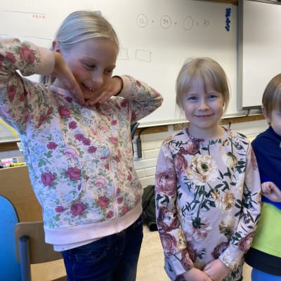 Kaksi kukkapaitaan pukeutunutta pientä tyttöä ja tummansiniseen pusakkaan pukeutunut pieni poika hymyilee ja poseeraa kameralle luokkahuoneessa.