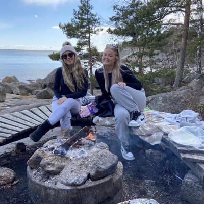 Pinja och Riina sitter och ler vid en öppen eld med havet i bakgrunden.