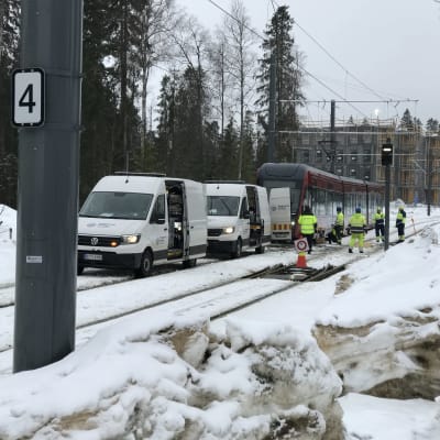 Kaksi valkoista pakettiautoa ja punainen raitiovaunu seisovat jonossa ratikkakiskojen kohdalla Tampereen Hervantajärvellä.