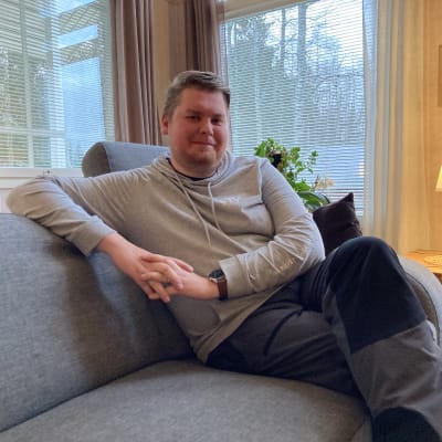 Mies istuu olohuoneen sohvalla katsoen kameraan.