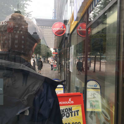 En man står utanför en R-kiosk och människor går på gatan omkring honom. Bilden är arrangerad.