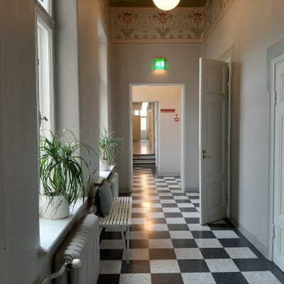 lång korridor med svartvitrutigt golv, vita spegeldörrar på gläntoch fönstersmygar med palmer i kruka