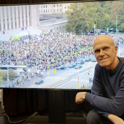 Thomas Wallgren i Svenska Yles lokaler framför en tv-skärm. På tv-bilden syns klimatdemonstranter utanför riksdagen den 27 september.