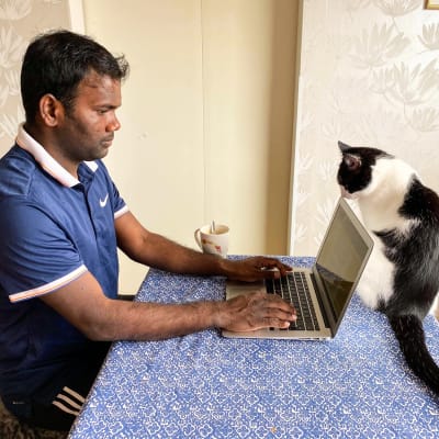 En man sitter vid köksbordet och skriver på en bärbar dator. En katt snusar på datorn.