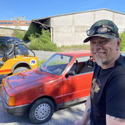 Man med skärmmössa framför liten röd bil.